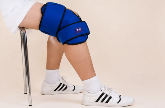 A person seated on a chair wearing a bright blue knee support brace on their right leg, with white socks and white Adidas sneakers, against a plain beige studio background.