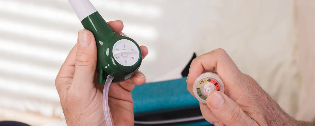 Hand holding a green and black device with a white nozzle, likely an inhaler, against a blurred indoor background.